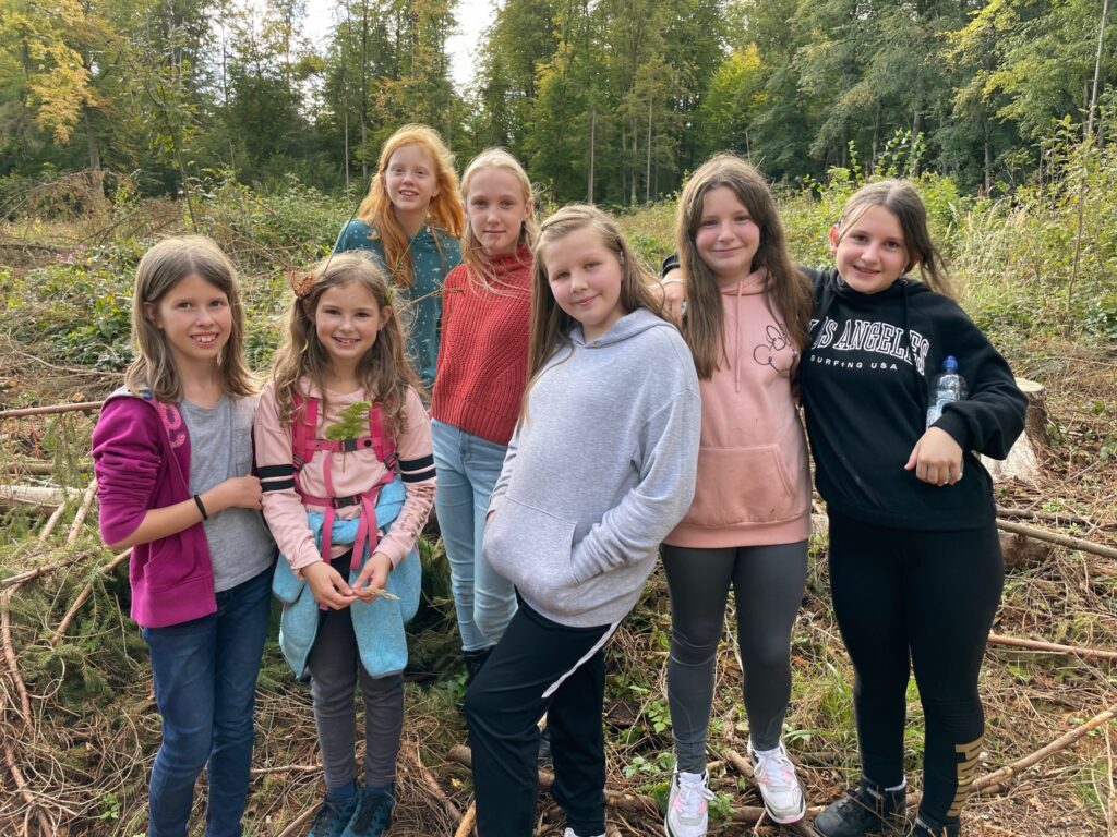 Gruppe von sieben Kindern steht im Freien, umgeben von Bäumen und Sträuchern.