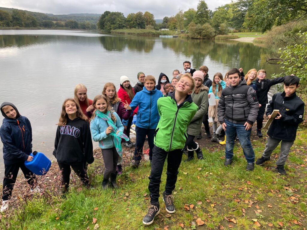 Schulklasse steht am Ufer eines Sees und posiert fröhlich für ein Foto.