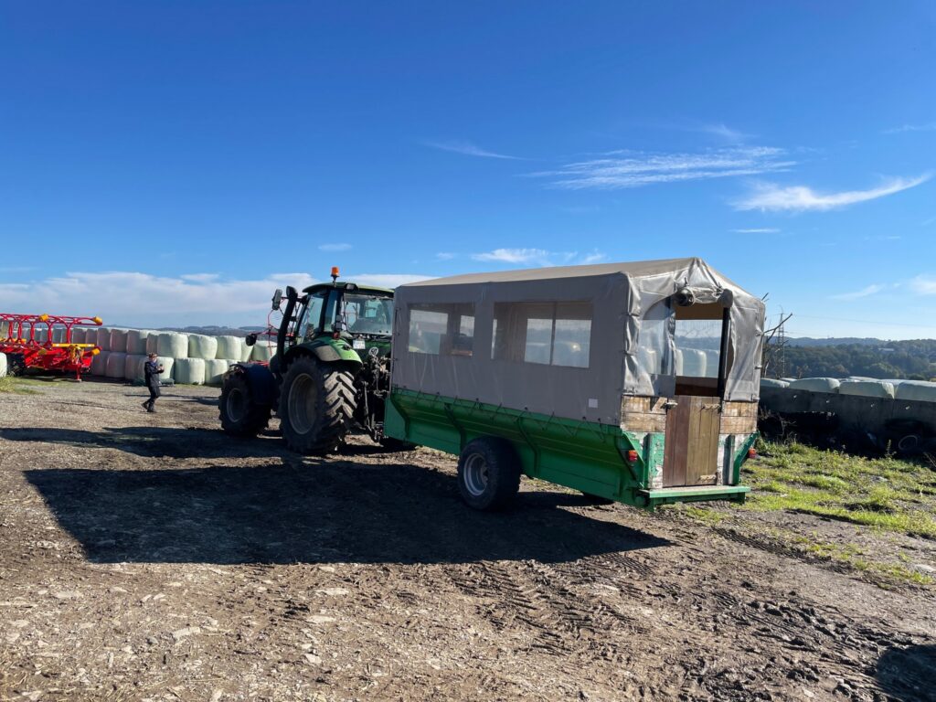 Traktor mit Anhänger auf einem Feld, im Hintergrund Heuballen. Klarer blauer Himmel.