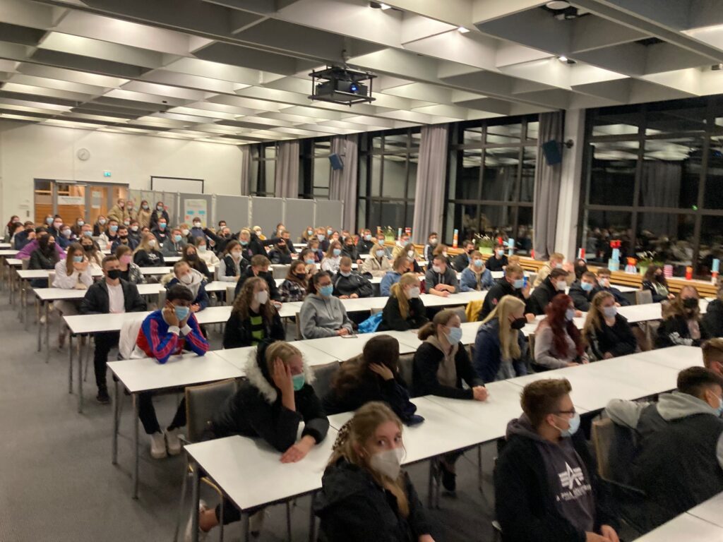 Schülerinnen und Schüler sitzen in einem großen Klassenraum an Tischen.
