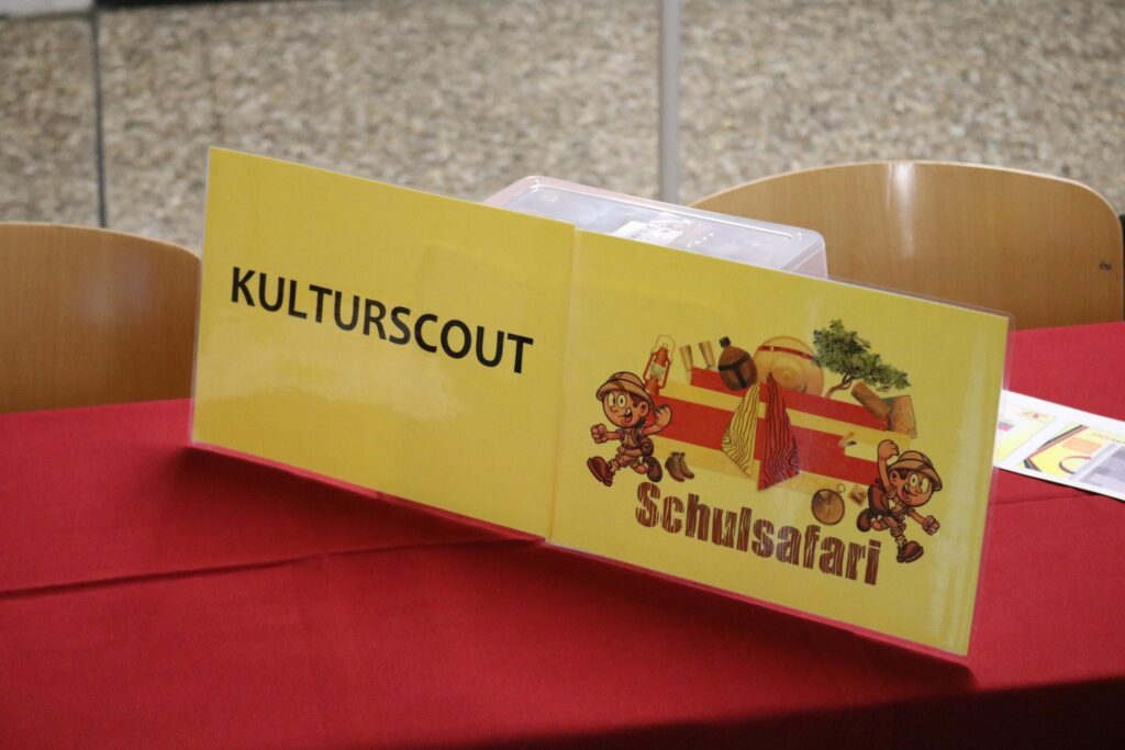 Tisch mit gelbem Schild „KULTURSCOUT“ und Logo „Schulsafari“.