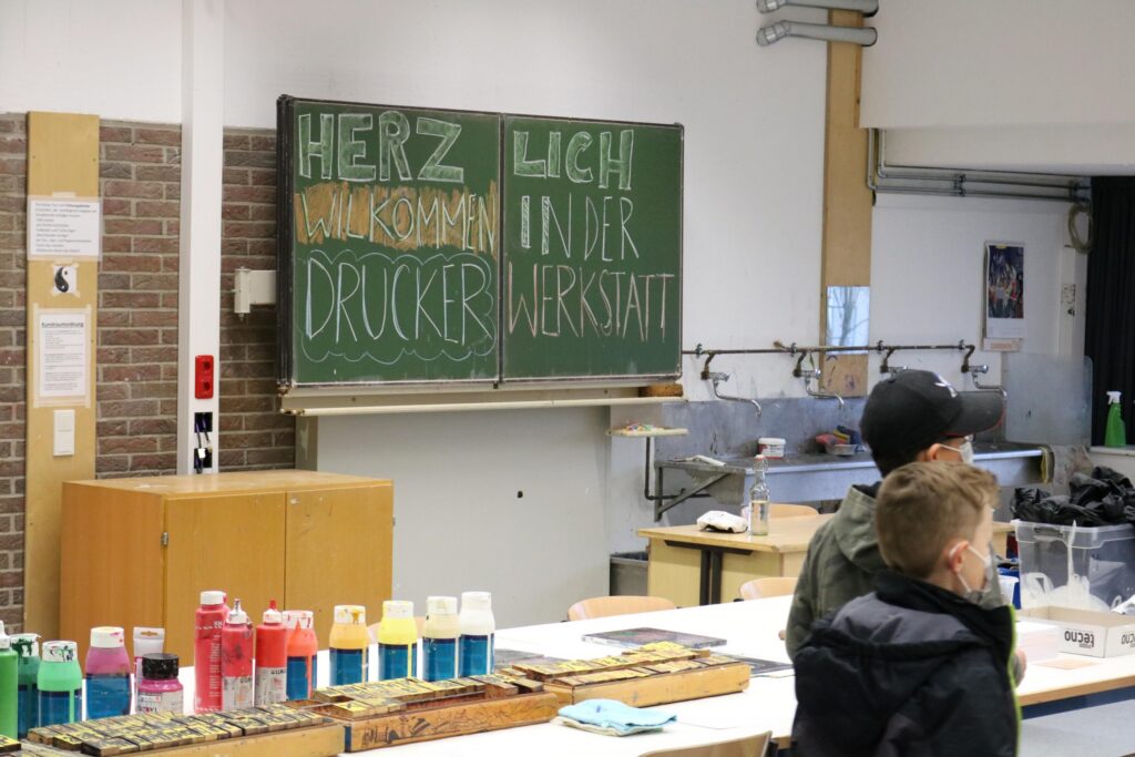 Grüne Tafel mit der Beschriftung "HERZLICH WILLKOMMEN IN DER DRUCKERWERKSTATT".
