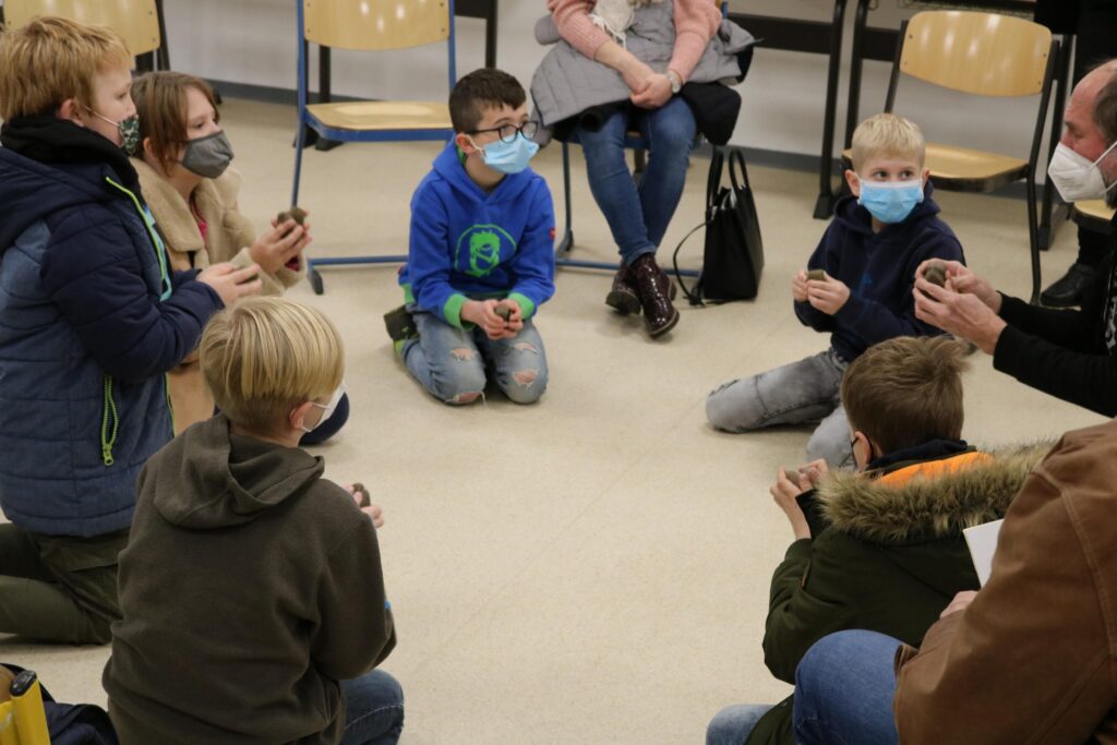 Kinder sitzen im Kreis, aktiv am Gruppenprojekt beteiligt. Einige tragen Masken.