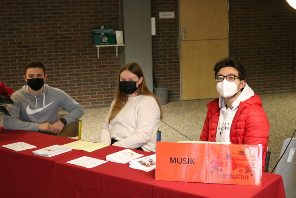 Drei Schüler an einem Tisch mit Musikinformationen und Masken.