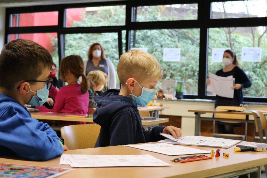 Schüler in Klassenraum mit Mund-Nasen-Schutz, Lehrerin erklärt am Pult.