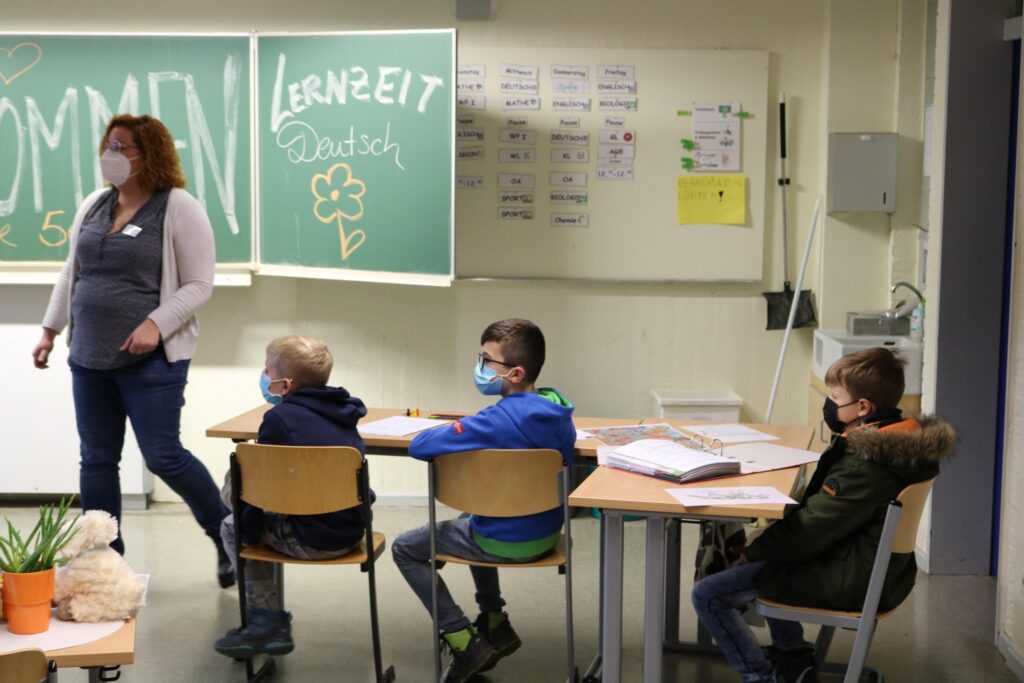 Lehrerin steht vor einer Klasse mit drei Schülern, alle tragen Masken.