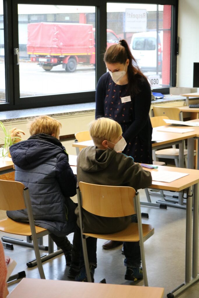 Lehrerin erklärt etwas an Schreibtischen mit zwei Schülern im Klassenzimmer.