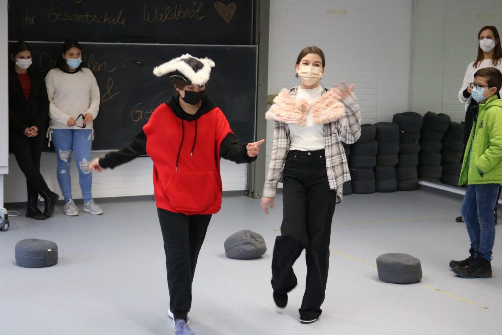 Zwei Mädchen in Kostümen führen ein kleines Theaterstück auf. Hintergrund mit Zuschauern.