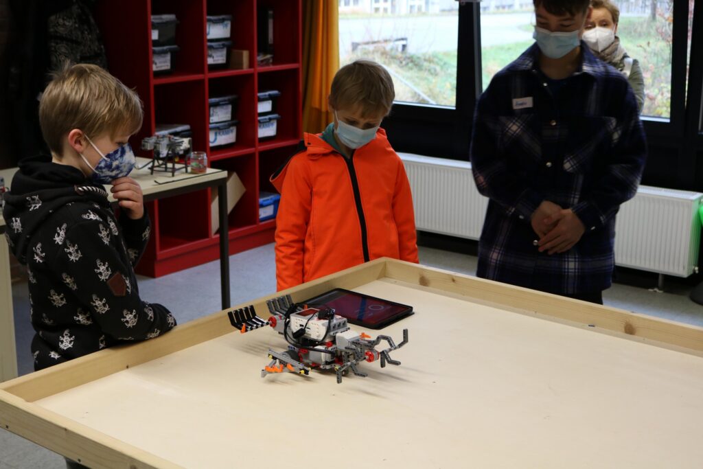 Kinder beobachten einen Roboter auf einem Tisch in einem Klassenraum.