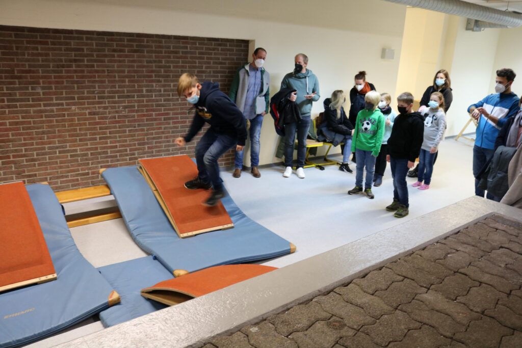 Schüler springt auf Turnmatten, Gruppe schaut zu, einige tragen Masken.