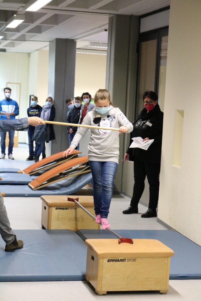 Schülerin balanciert auf einer Slackline zwischen zwei Kästen. Zuschauer beobachten.