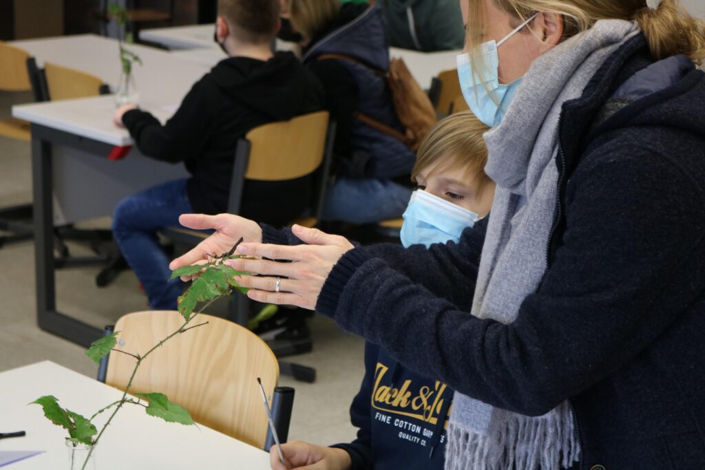 Lehrerin zeigt Schülern eine Pflanze im Klassenraum. Schüler tragen Maske.
