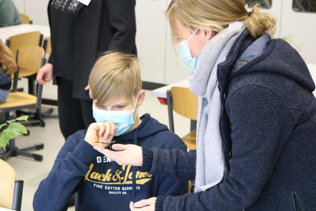 Schüler mit Maske bei der Arbeit, Lehrerin zeigt ihm etwas.