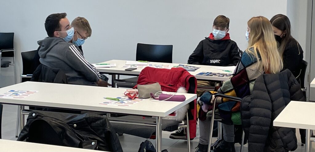 Schüler und Lehrer sitzen an einem Tisch, tragen Masken und arbeiten zusammen.