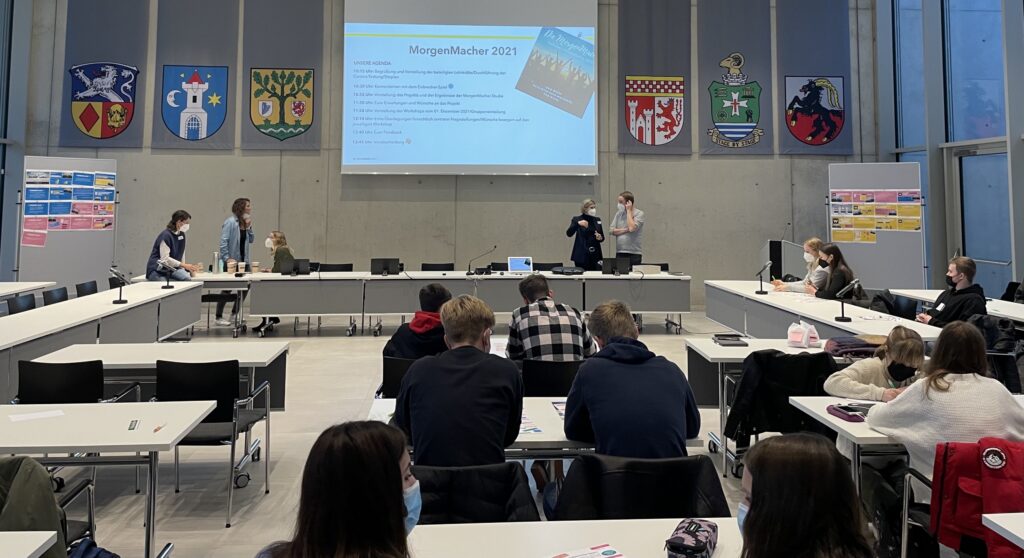 Präsentation in einem großen Raum mit Tischen und Plakaten an der Wand.