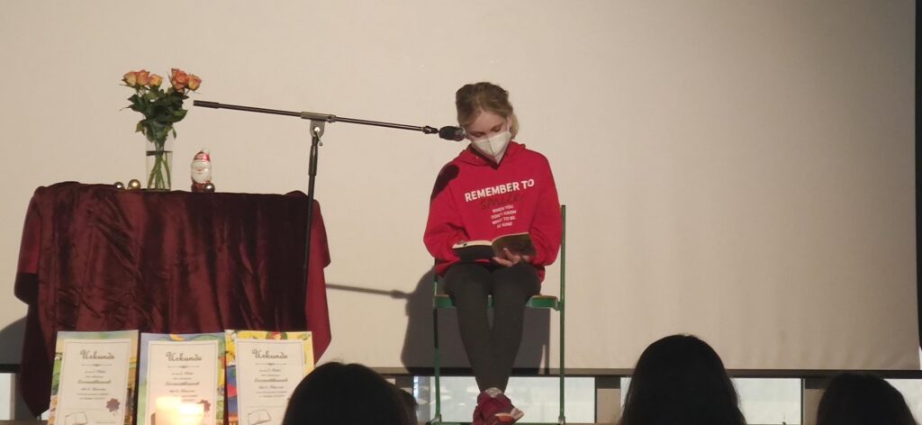 Ein Mädchen sitzt auf einem Stuhl und liest aus einem Buch vor.