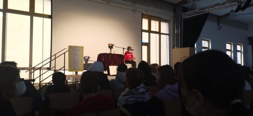 Schüler sitzt auf der Bühne vor Publikum, Blumen auf dem Tisch.