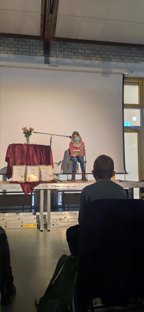 Eine Person sitzt an einem Tisch vor einem Publikum in einem Saal.