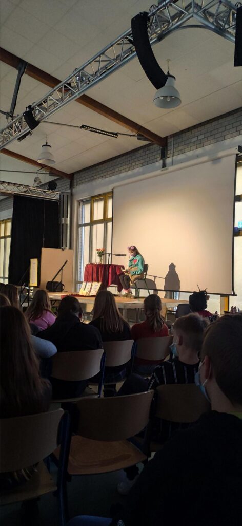 Eine Person steht auf einer Bühne vor Publikum, neben einem Tisch mit rotem Tuch.