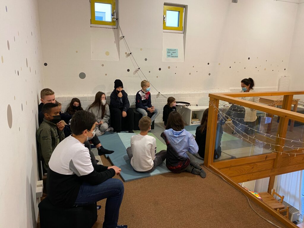 Gruppe von Schülern sitzt auf dem Boden und hört einer Lehrkraft zu.