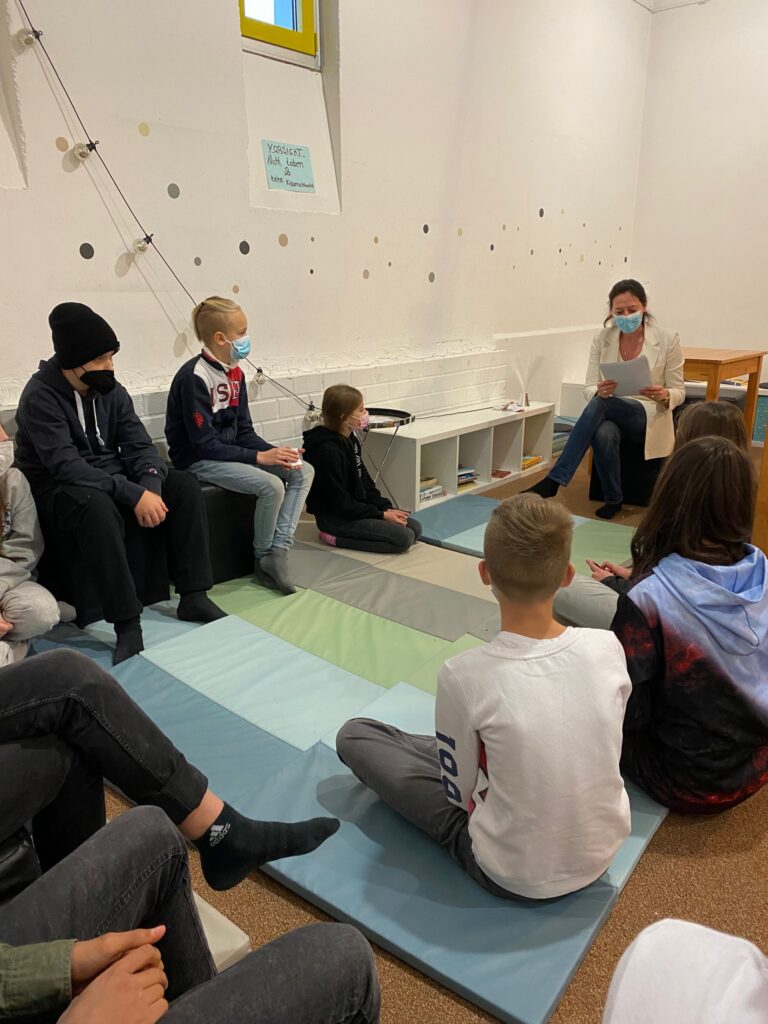 Schüler sitzen auf Matten und hören einer Lehrkraft während einer Unterrichtsstunde zu.