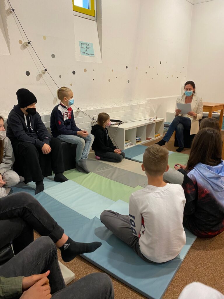 Schüler sitzen im Kreise und hören einer Lehrerin zu.