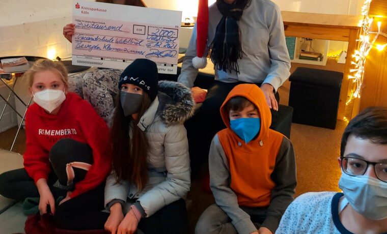 Gruppe von sechs Personen mit Masken, Weihnachtsmütze und Kerzen in einem Raum.