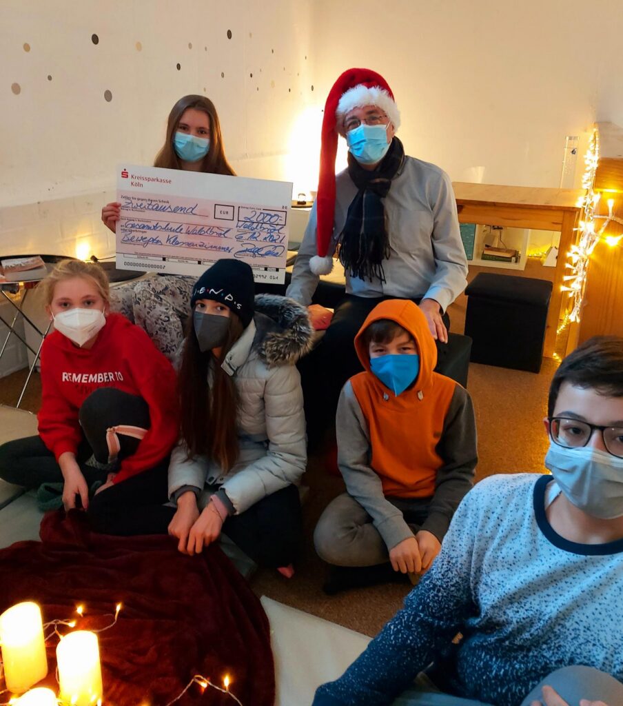 Gruppe von sechs Personen mit Masken, Weihnachtsmütze und Kerzen in einem Raum.