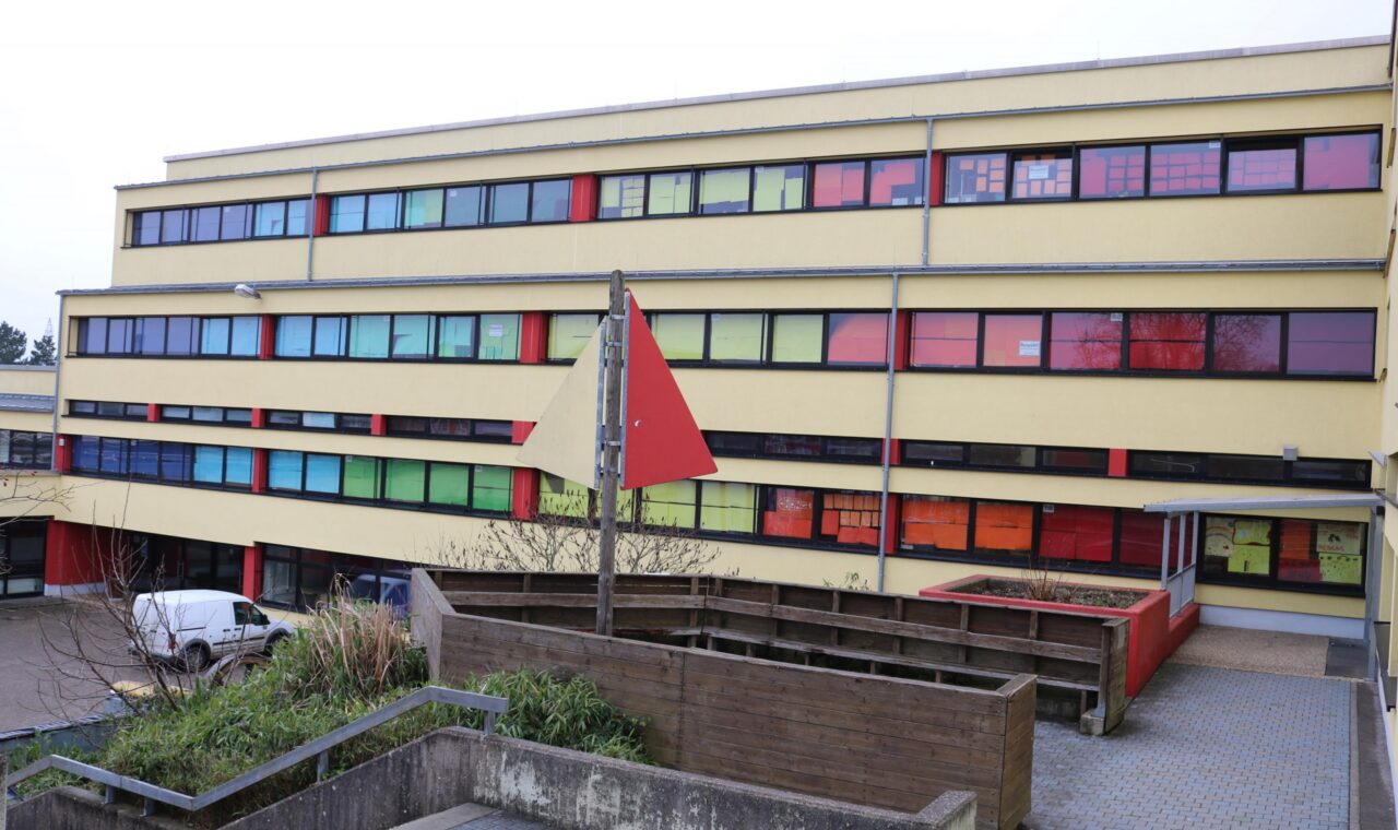 Schulgebäude mit bunten Fenstern und roter Fahne im Vordergrund.