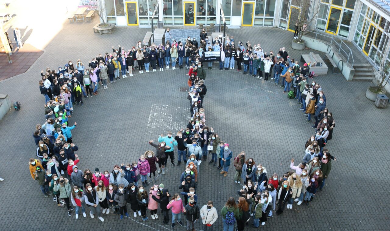Schüler stehen im Kreis und bilden ein großes Friedenssymbol auf dem Schulhof.