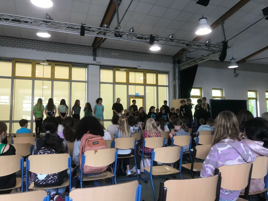 Schülerinnen und Schüler stehen auf der Bühne vor einer sitzenden Gruppe.
