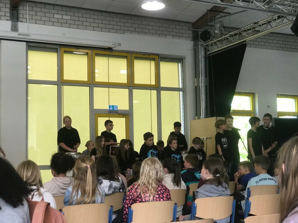 Schüler stehen auf einer Bühne vor einer Publikumssitzung in einer Aula.