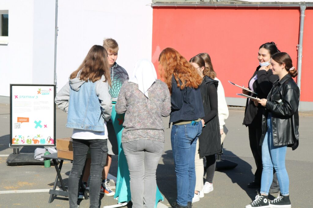 Gruppe von Schülern diskutiert im Freien, farbige Wände im Hintergrund.