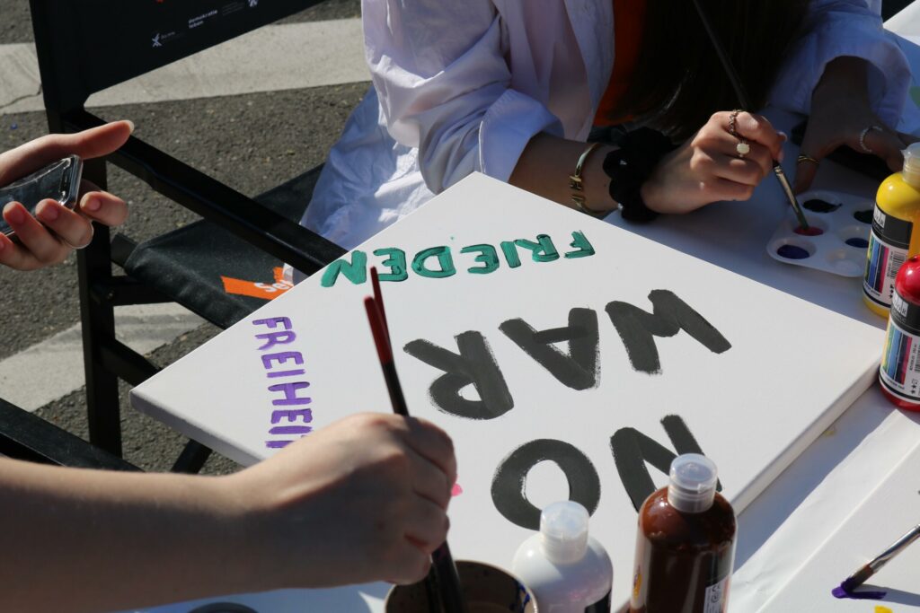 Hände halten Pinsel und bemalen ein Schild mit Worten wie „NO WAR“ und „FREIHEIT“.