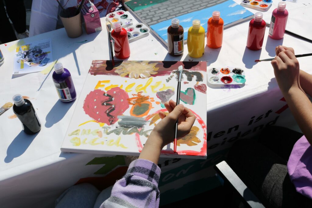 Malende Hand mit Pinsel über einem bunten Kunstwerk auf einem Tisch. Farben in Bechern daneben.