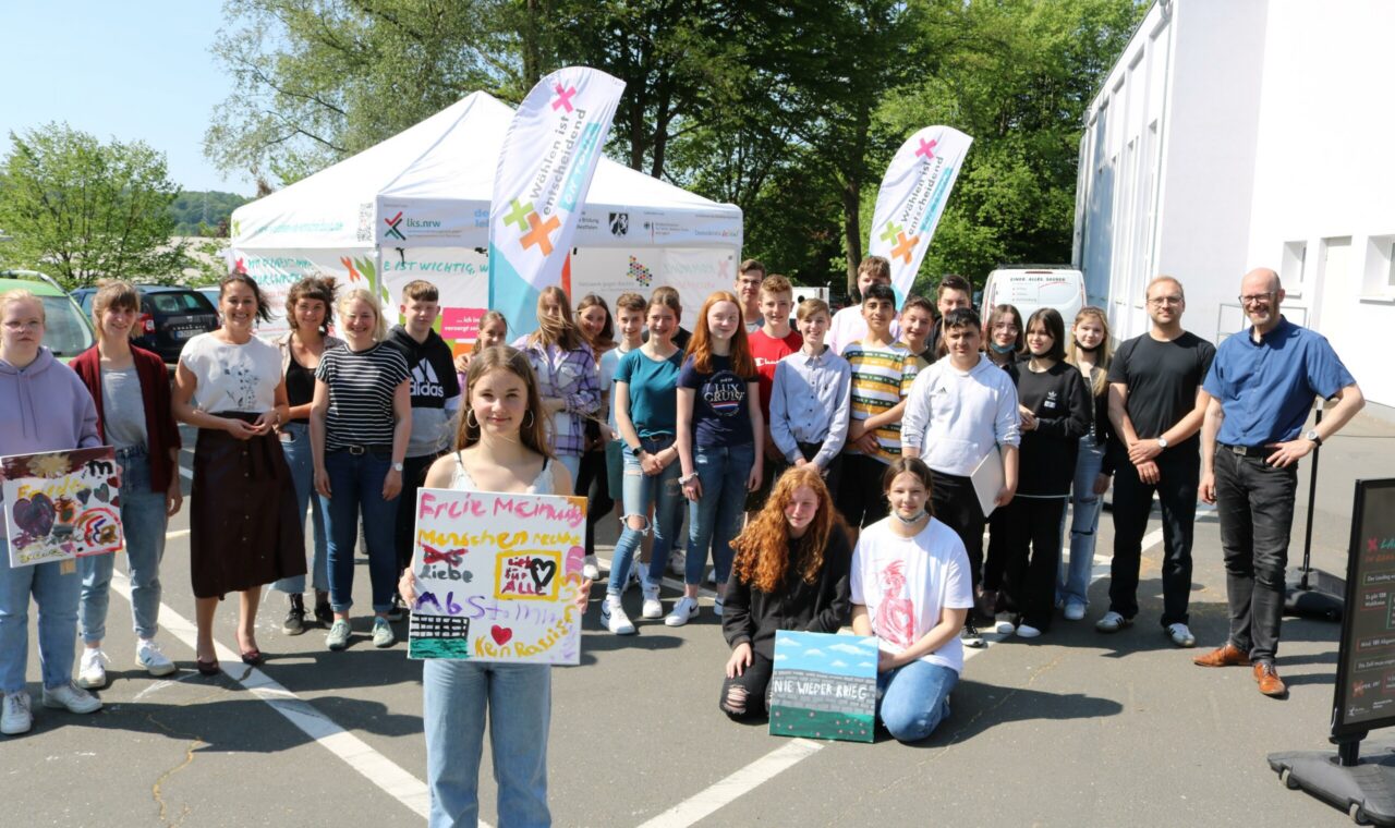 Gruppe von Schülern und Lehrern vor einem Zelt mit Bannern, lächelnd und posierend.