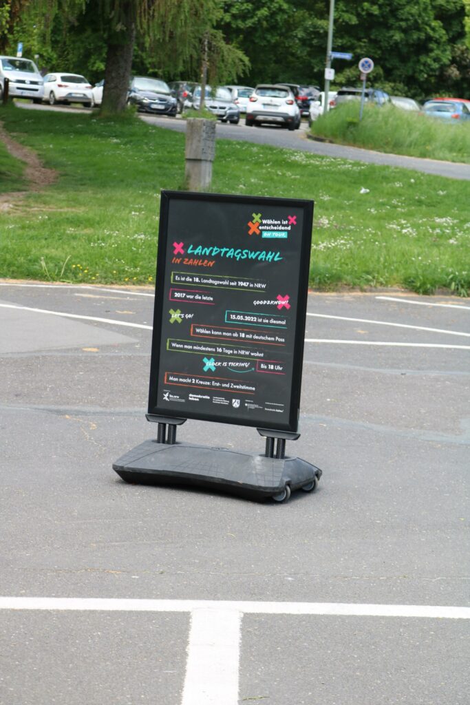 Informationstafel mit Veranstaltungshinweisen auf einem Parkplatz.