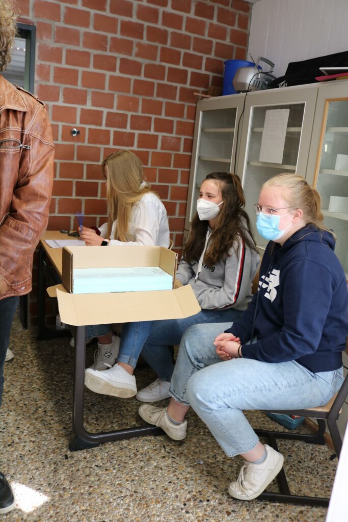 Drei Schülerinnen sitzen an einem Tisch, tragen Masken und sind in einem Klassenraum.