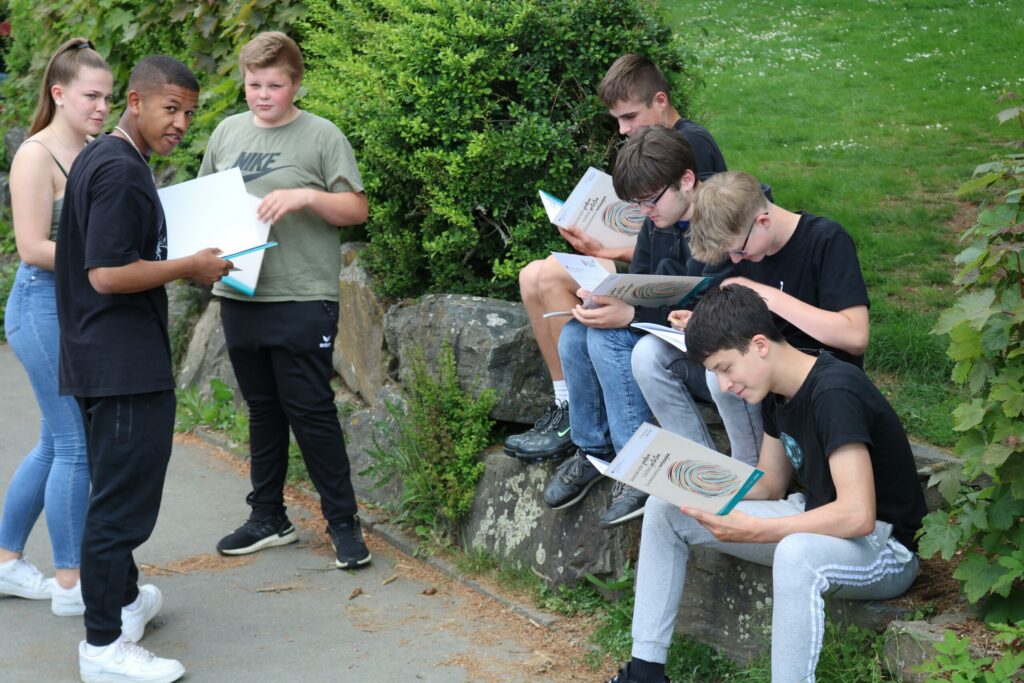 Gruppe von Schülern sitzt und steht im Freien mit Unterlagen in der Hand.