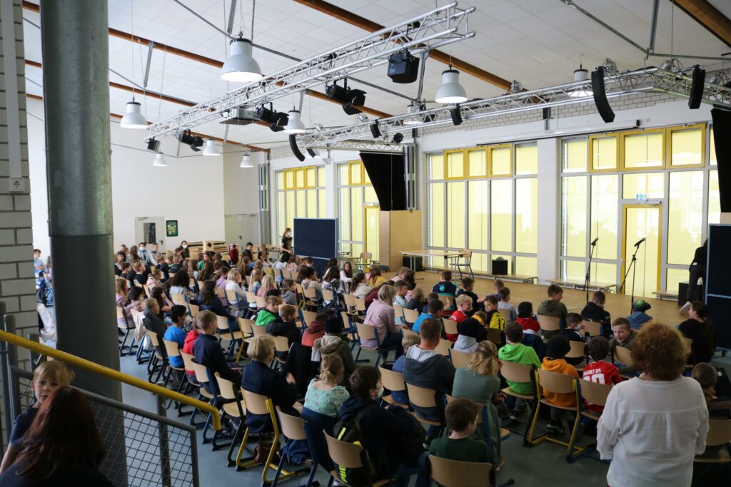Schüler sitzen in einem großen Raum, Bühne im Hintergrund, gut beleuchtet.