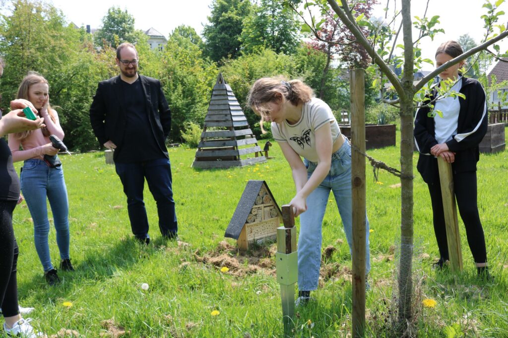 Schüler pflanzen einen Baum auf einer Wiese, Zuschauer beobachten.