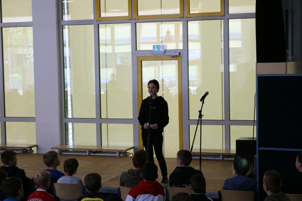 Ein Schüler steht auf einer Bühne und spricht vor einer Gruppe von Mitschülern.