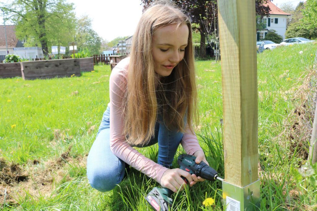 Mädchen mit Bohrmaschine befestigt einen Pfahl im Garten.