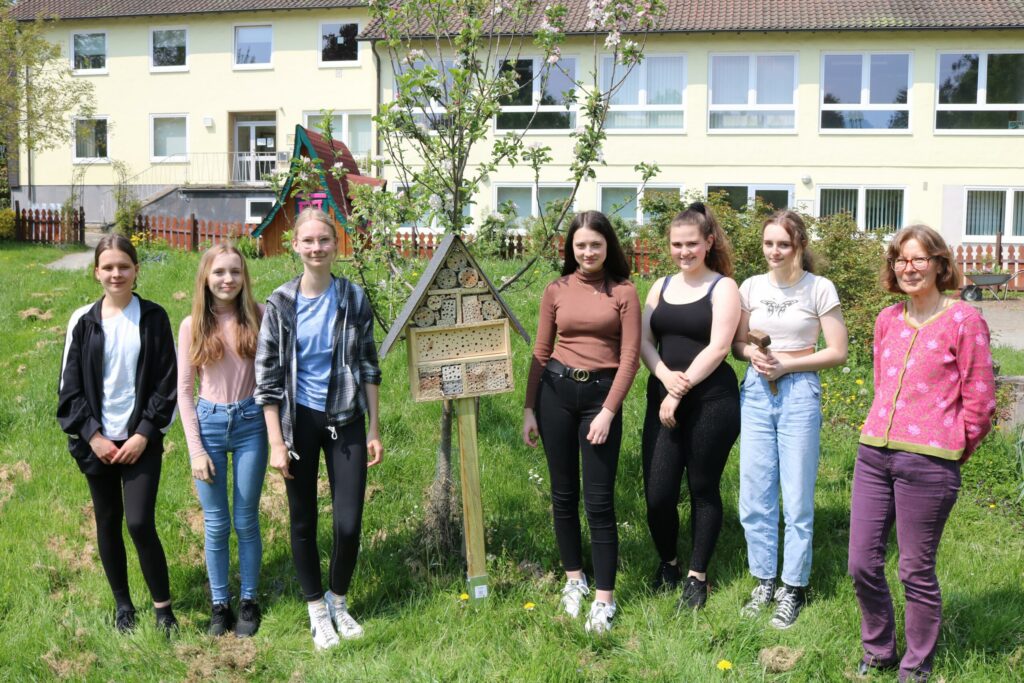Sechs Schülerinnen und eine Lehrerin stehen lächelnd vor einem Baum und einem Schild im Schulhof.
