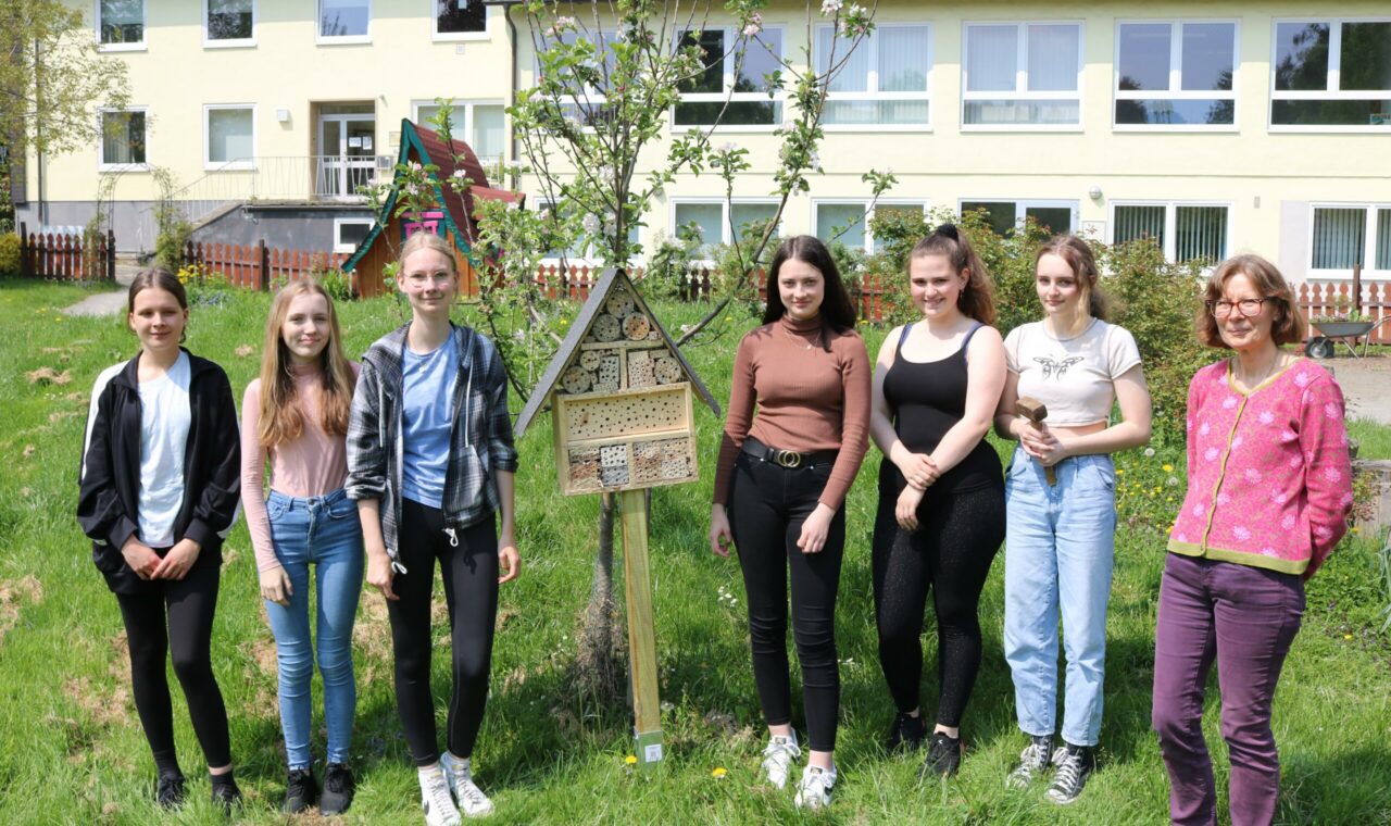 Sechs Schülerinnen und eine Lehrerin stehen lächelnd vor einem Baum und einem Schild im Schulhof.