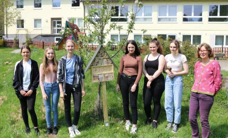 Sechs Schülerinnen und eine Lehrerin stehen lächelnd vor einem Baum und einem Schild im Schulhof.