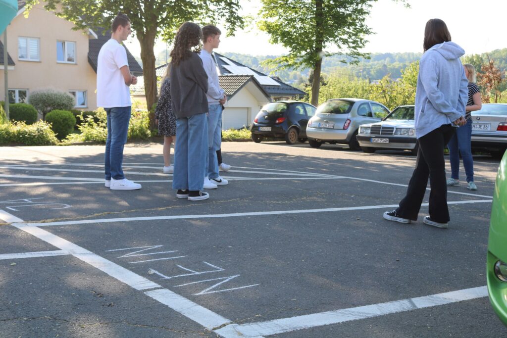 Gruppe von Jugendlichen steht auf einem Parkplatz, umgeben von Autos.