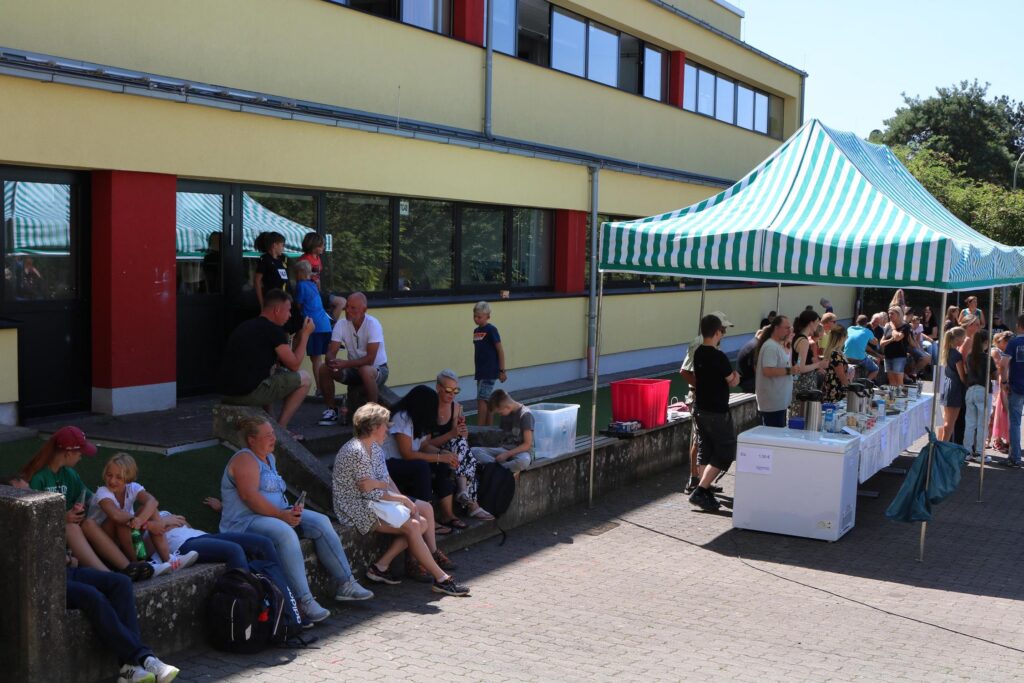 Schüler und Eltern sitzen entspannt vor einer Schule mit einem Festzelt.