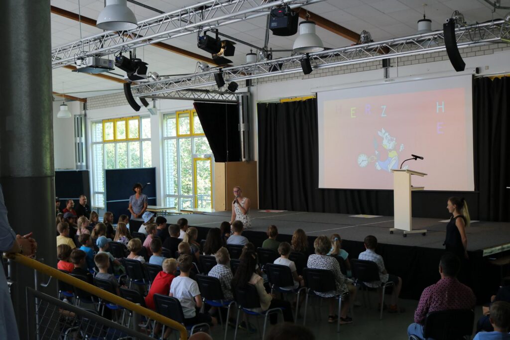Schulveranstaltung mit Publikum vor einer Bühne und Leinwand.