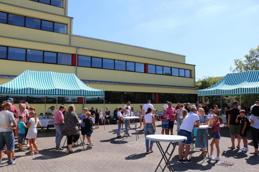 Viele Menschen stehen an Ständen vor einem Schulgebäude im Freien.
